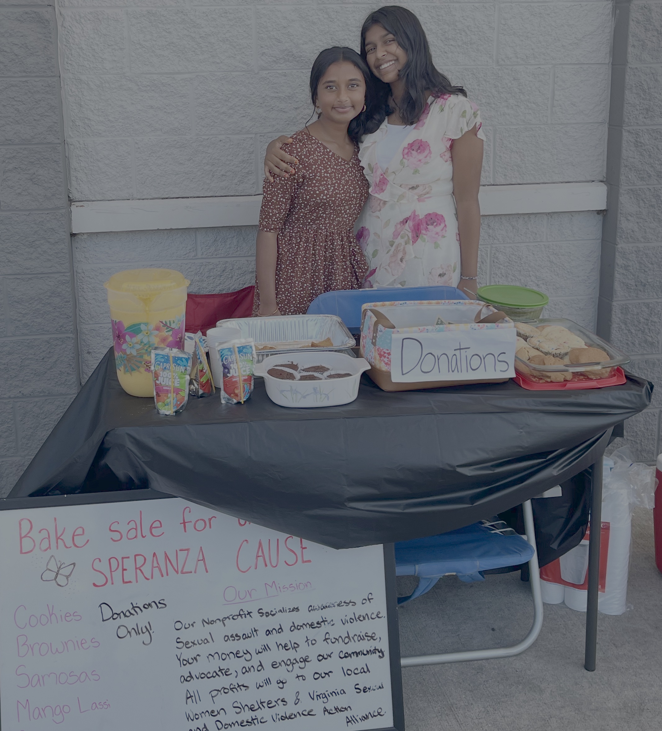 Two student leaders standing behind a Speranza Cause bake sale table with donations and homemade treats.